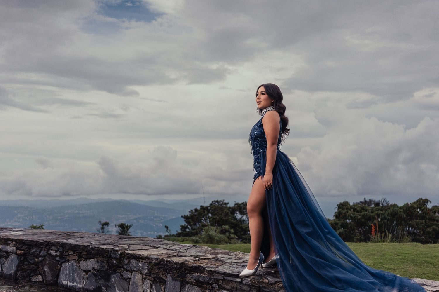 A person in a flowing blue gown stands against dramatic cloudy skies on an outdoor terrace or rooftop.
