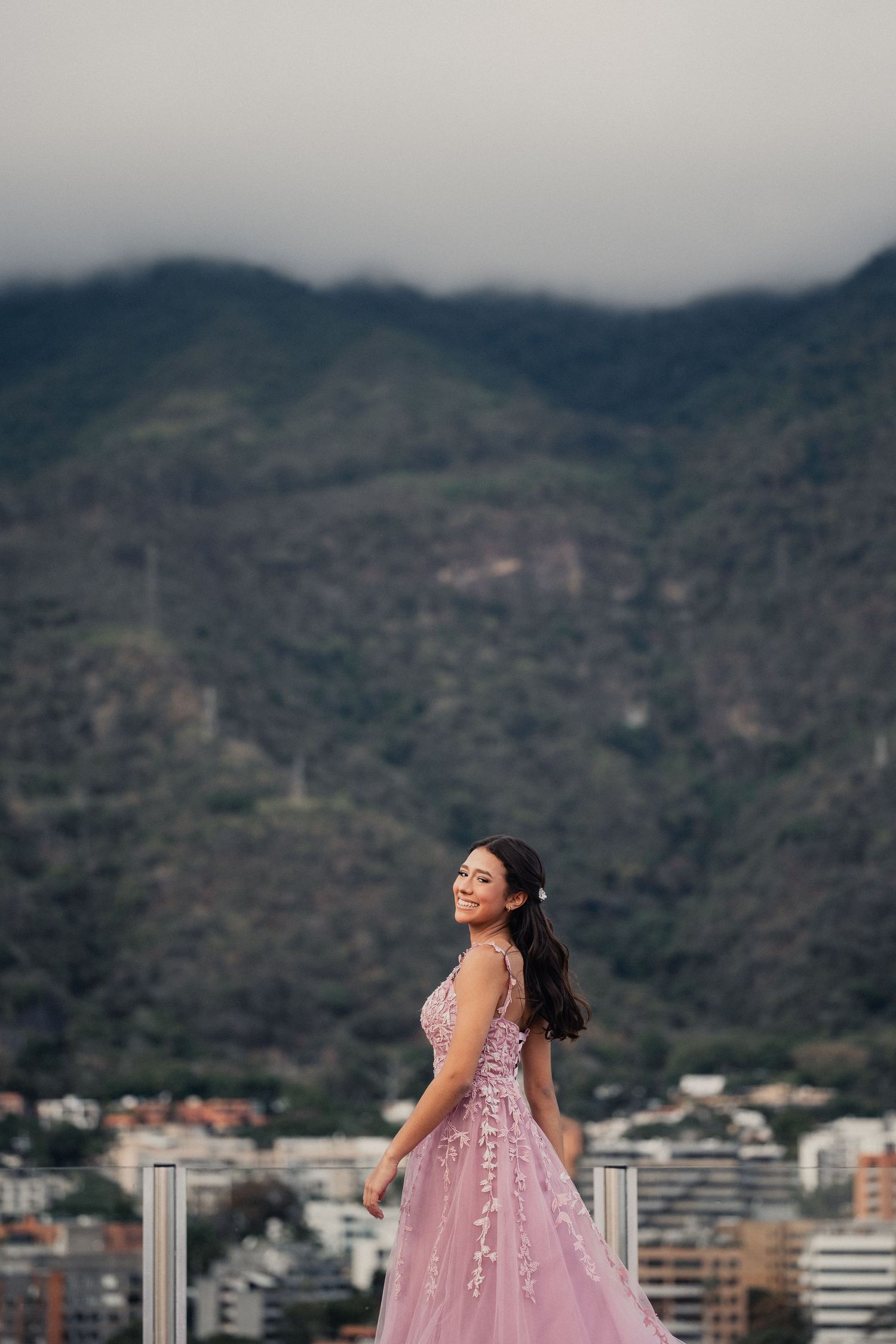 A person wearing a pink embellished gown poses on a rooftop with mountains and city buildings in the background.