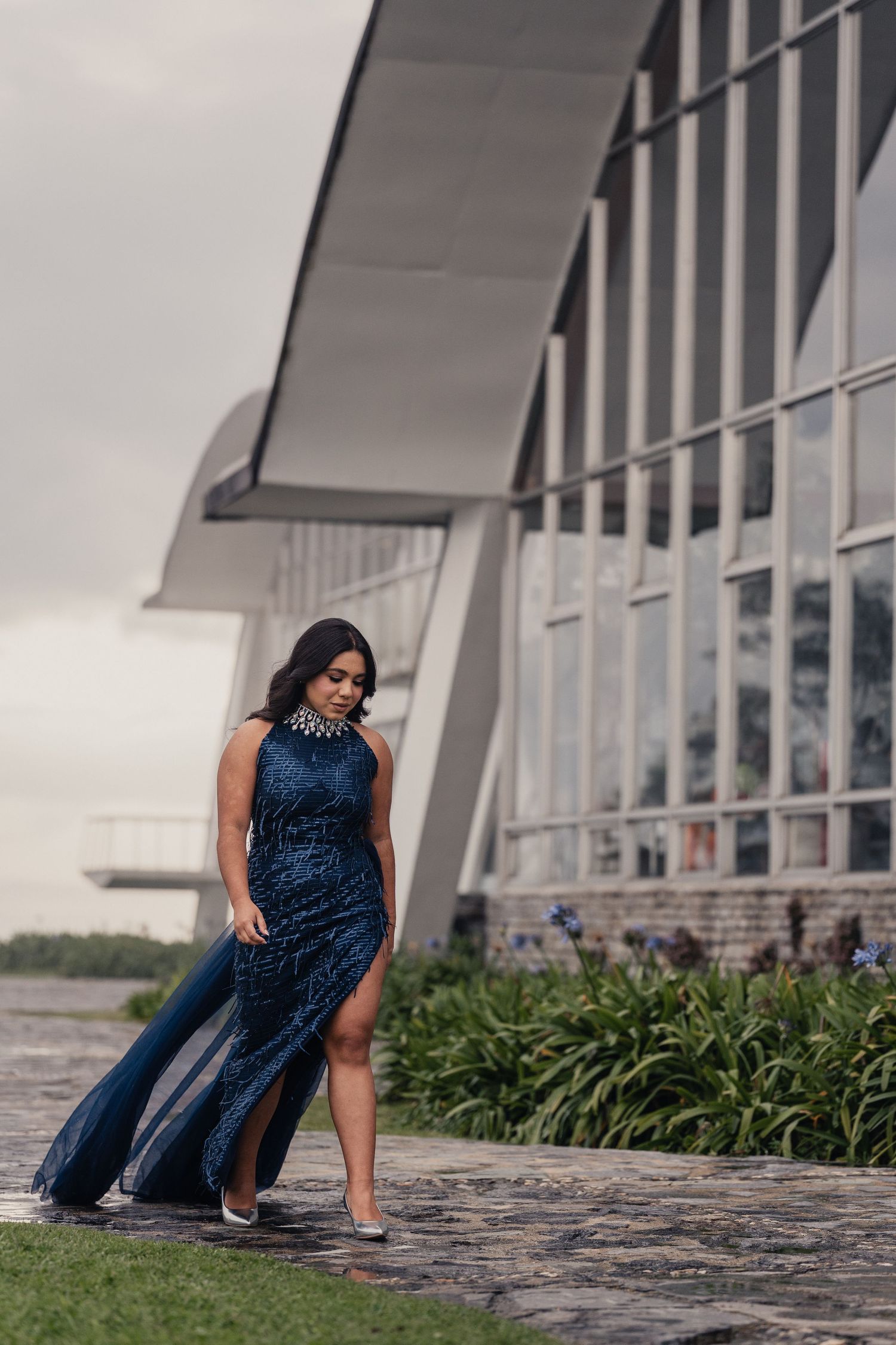 A woman in a blue dress poses gracefully on stone steps near modern architecture and landscaping.