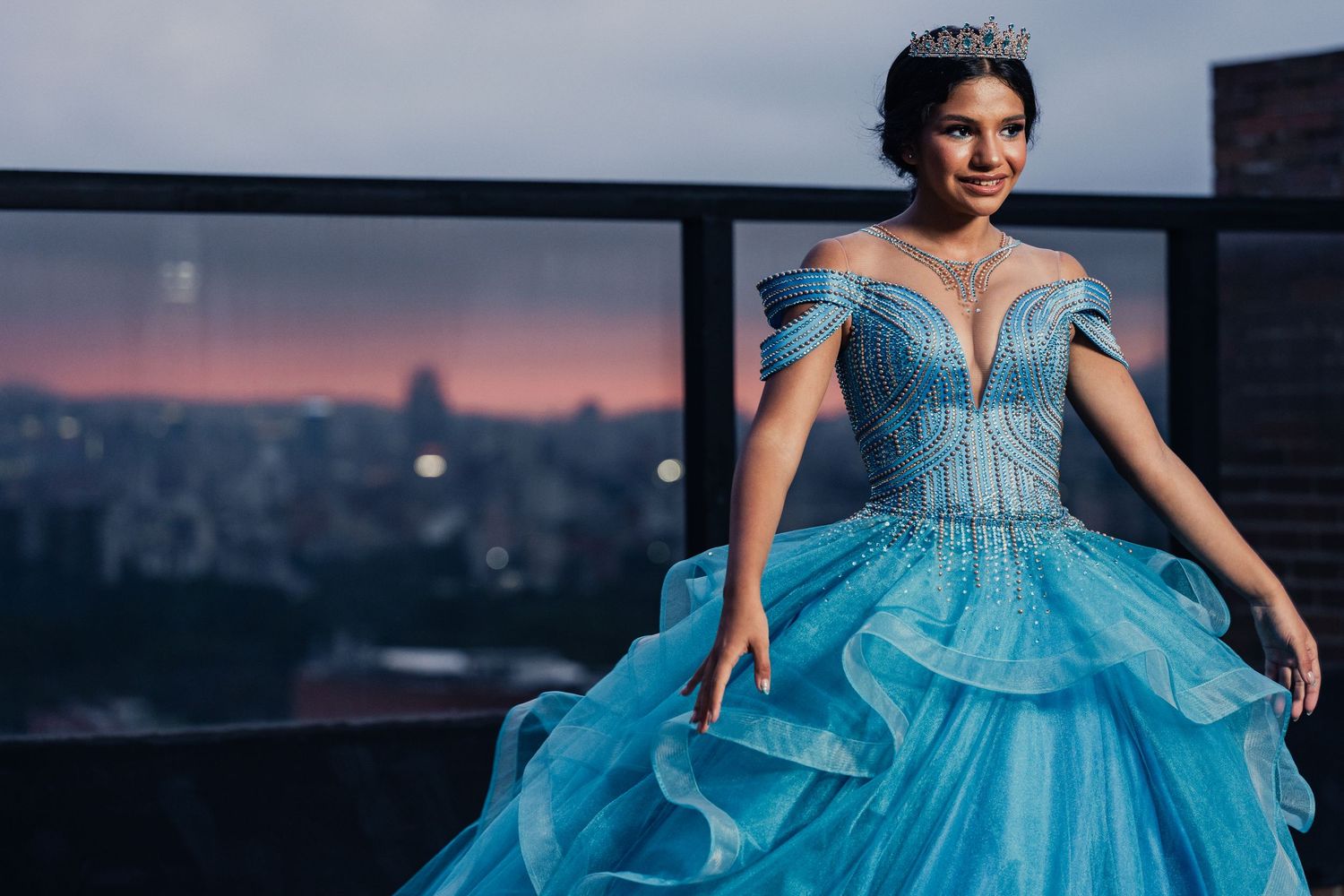 A stunning turquoise ball gown with crystal embellishments sparkles against a twilight cityscape backdrop.