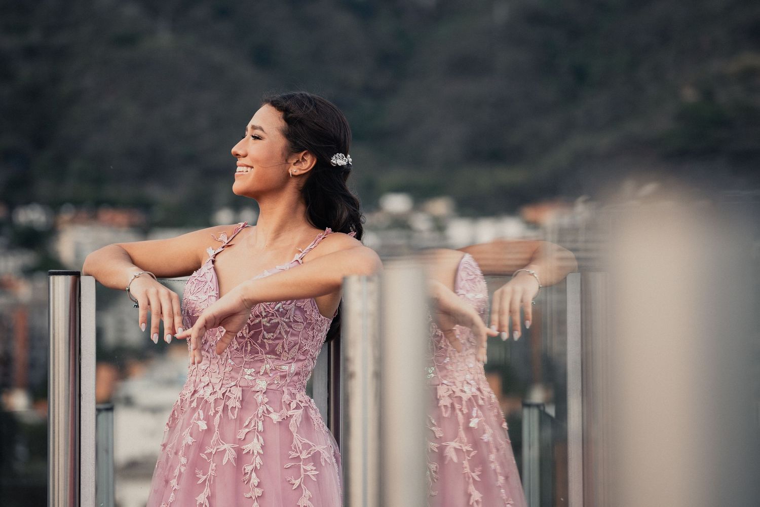 A woman in a pink lace dress leans against a balcony railing with mountains and buildings in the background.