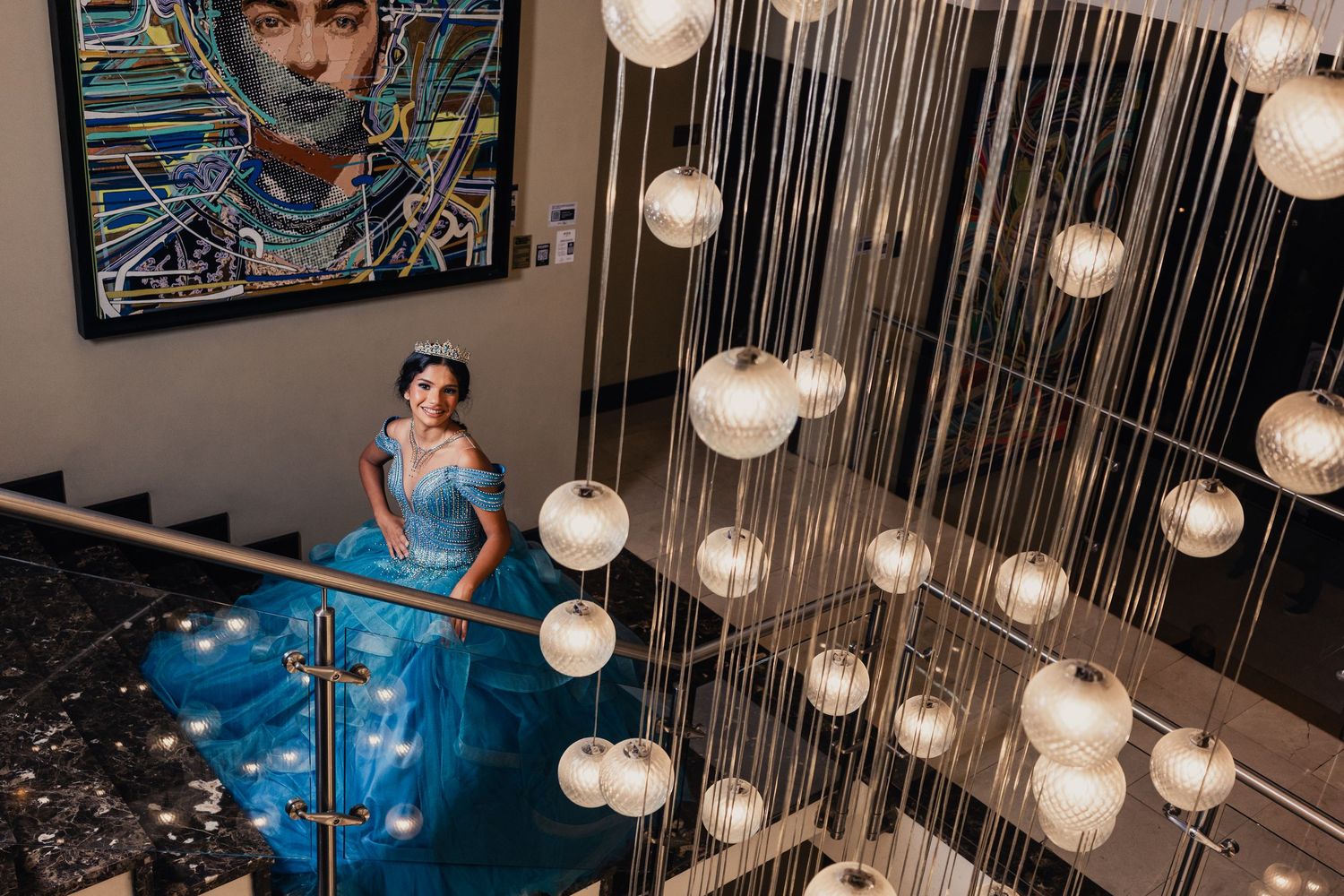 A person in a flowing blue gown stands on stairs beneath a modern chandelier with glowing orb lights and artistic wall decor.