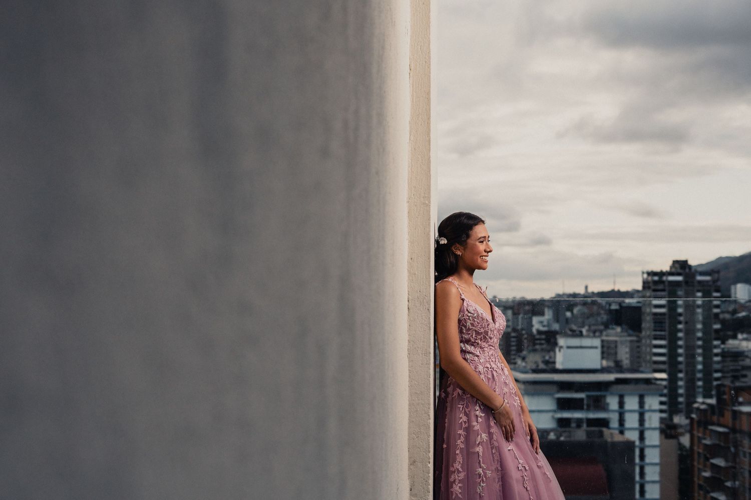 Woman in pink dress standing against urban backdrop with modern buildings and cloudy sky.