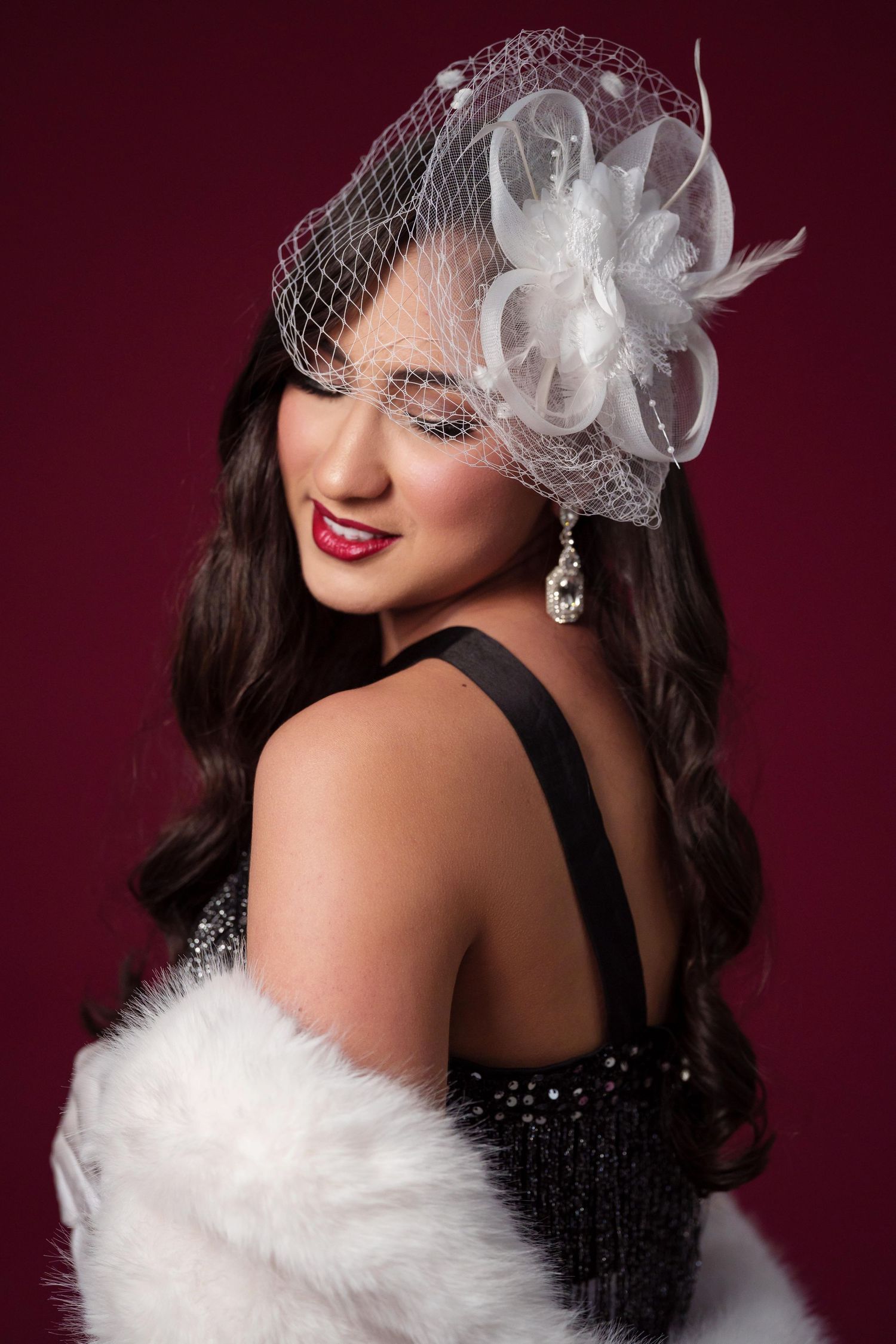 Glamorous vintage-inspired portrait featuring white feather fascinator, black sequin dress and white fur wrap against burgundy.