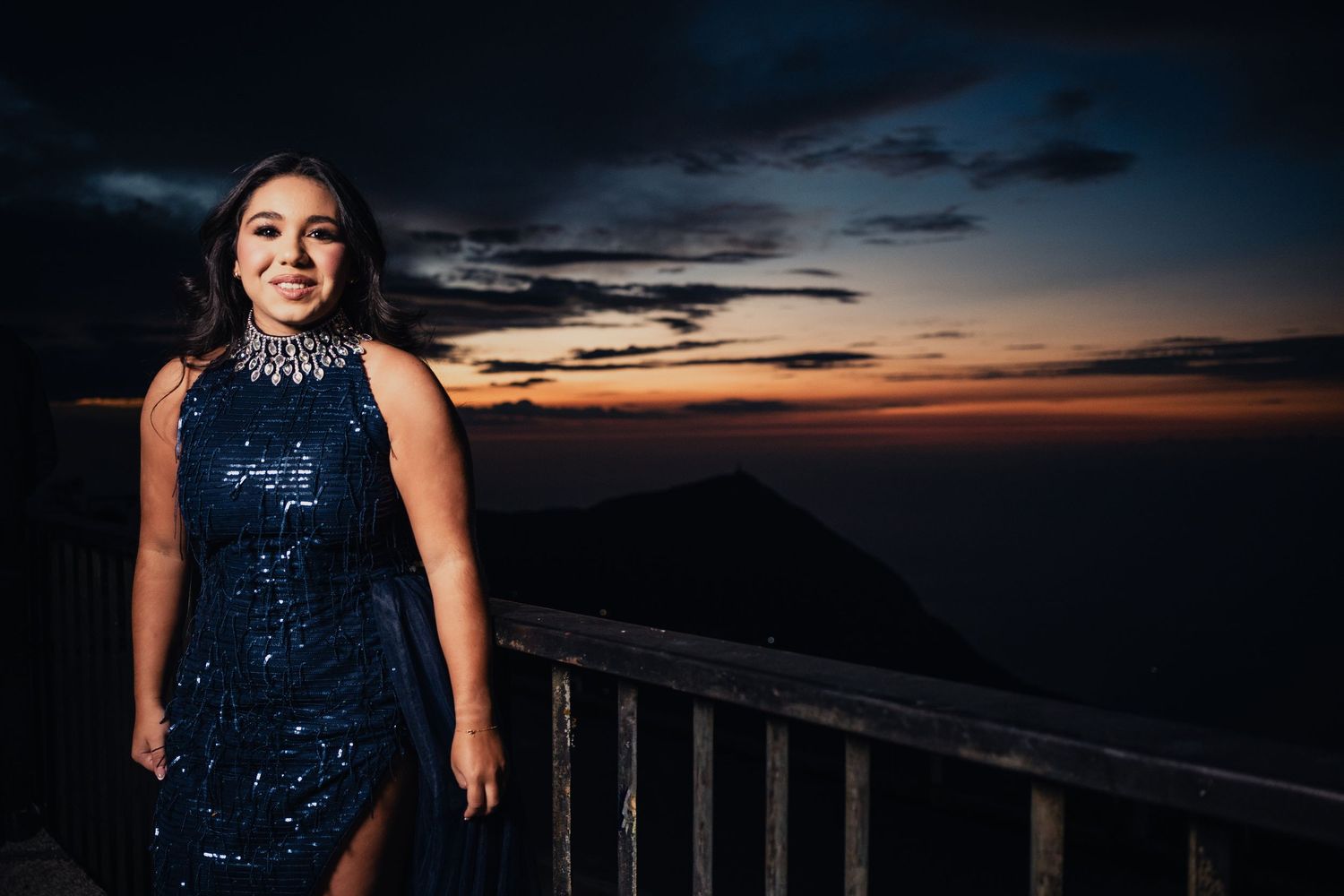 A person in a sequined dress poses against a dramatic sunset sky at a scenic overlook.