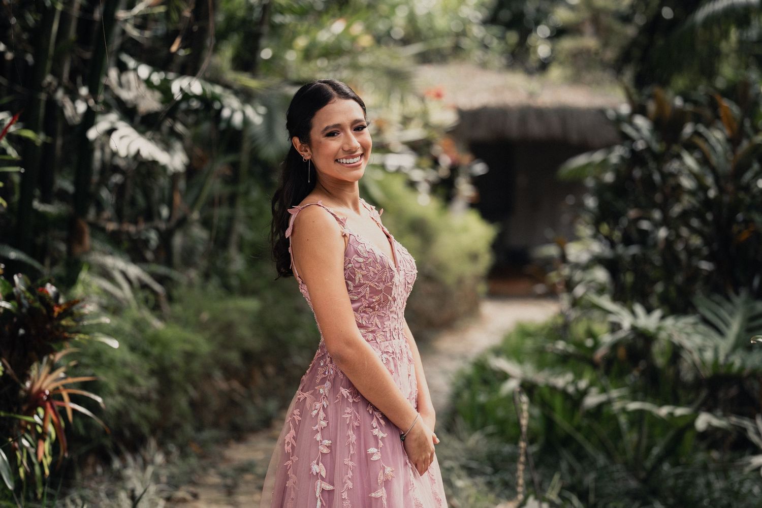 Woman in a pink lace dress poses in a lush garden setting with tropical plants and natural lighting.