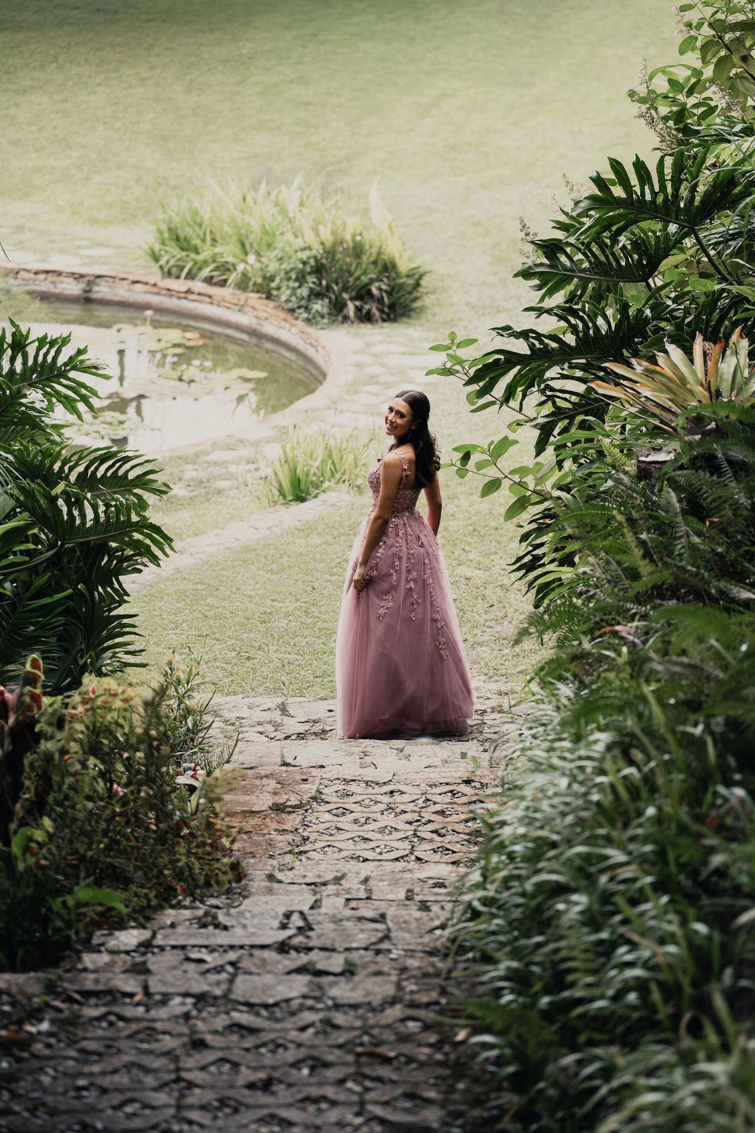 A figure in a dusty rose dress walks along a stone path surrounded by lush tropical foliage in a garden setting.
