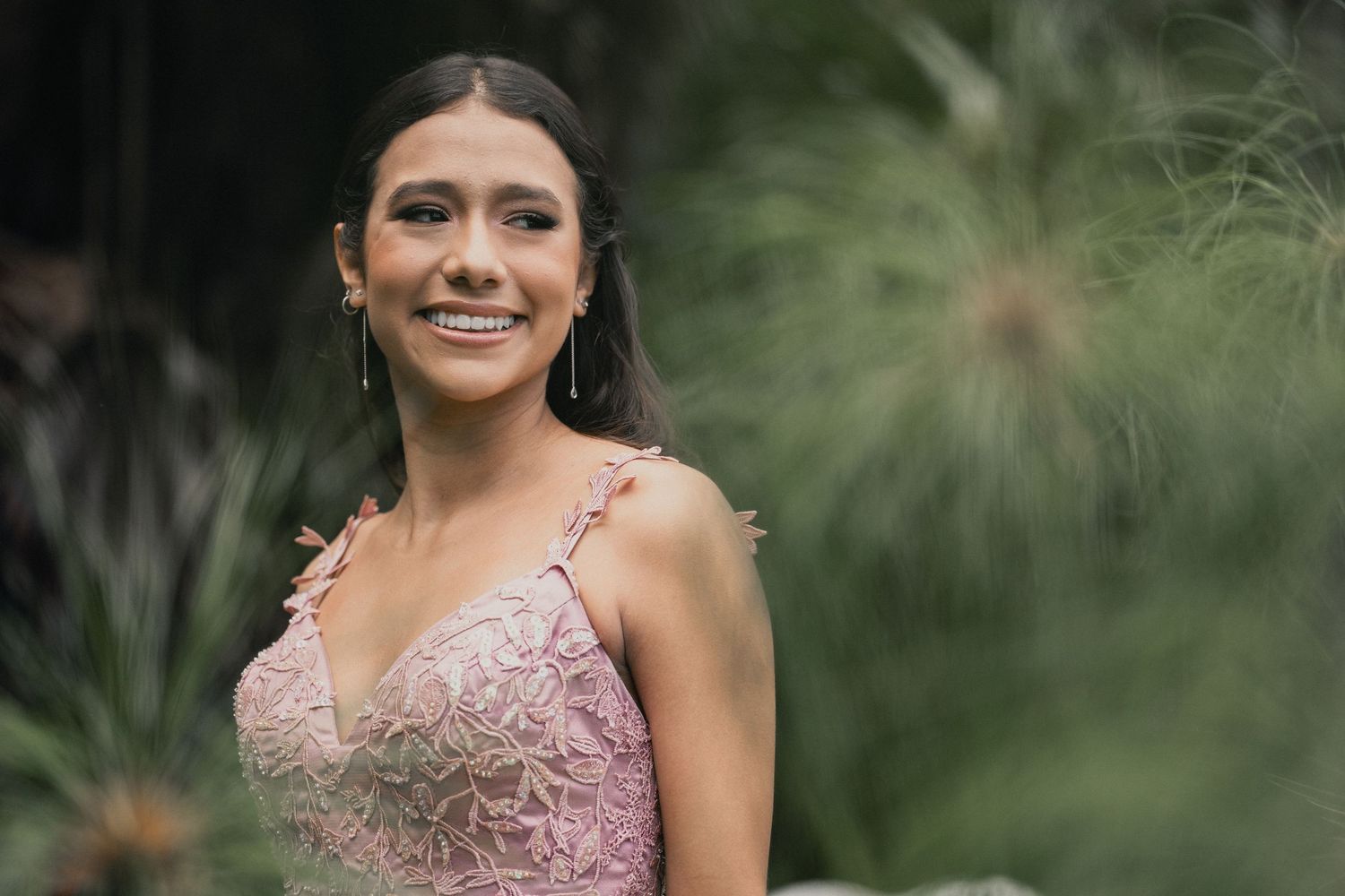 A person in a pink lace formal gown smiles while standing in front of soft-focus greenery outdoors.