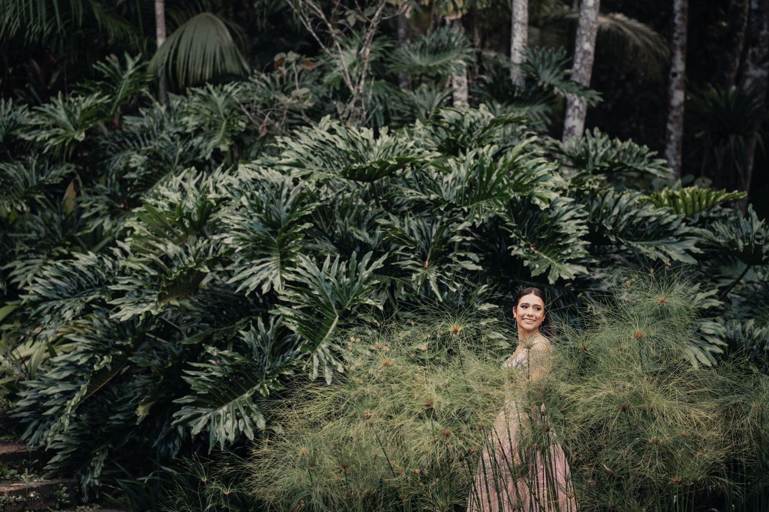 Woman in brown dress poses thoughtfully in a lush tropical garden setting with green foliage backdrop.
