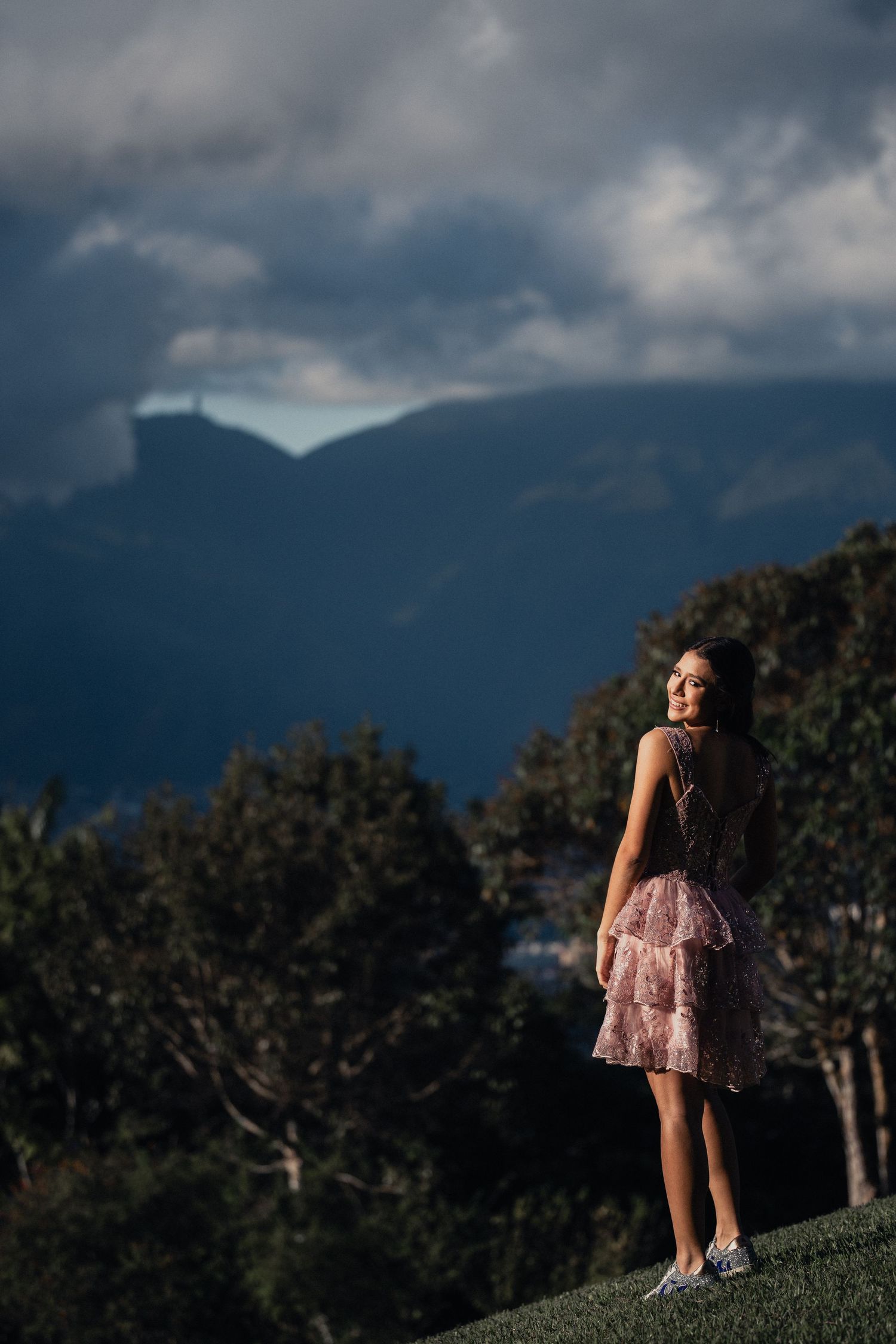 A person in a pink dress stands on a grassy hillside against a dramatic cloudy mountain backdrop at sunset.