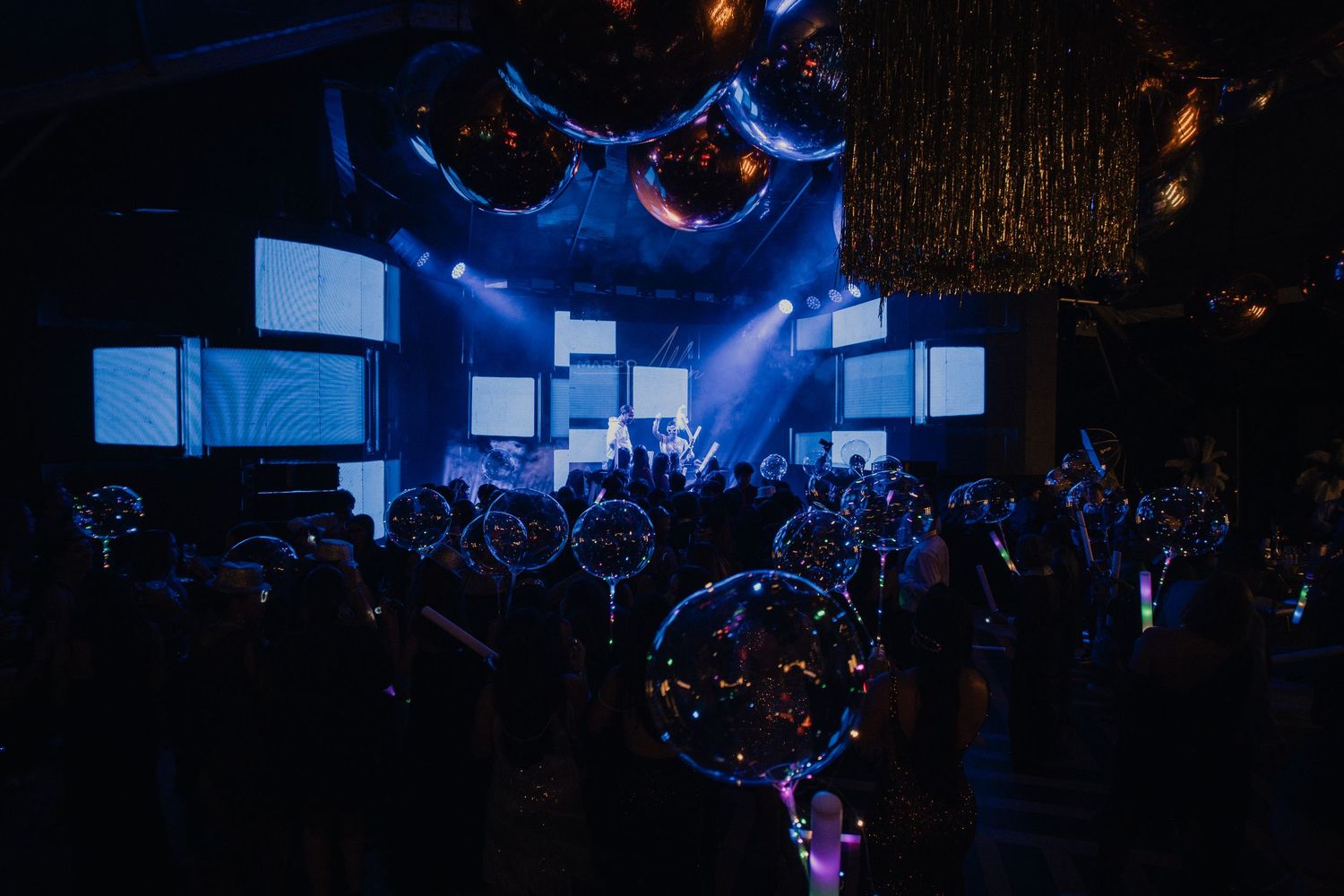 Blue spotlights illuminate multiple screens above a crowded dance floor with floating bubbles.