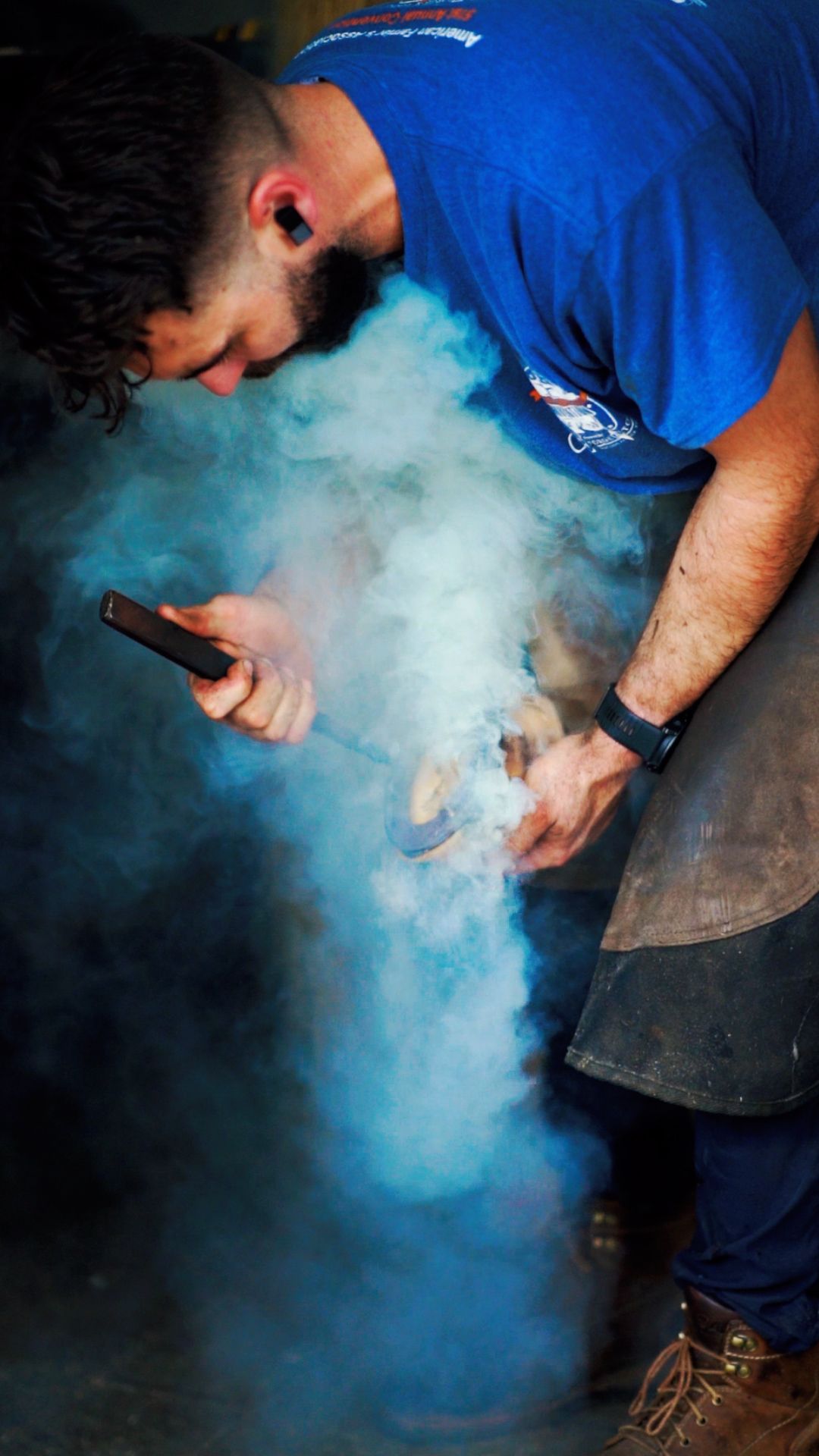 A person in a blue shirt works with blue smoke effects while holding tools against a dark background.