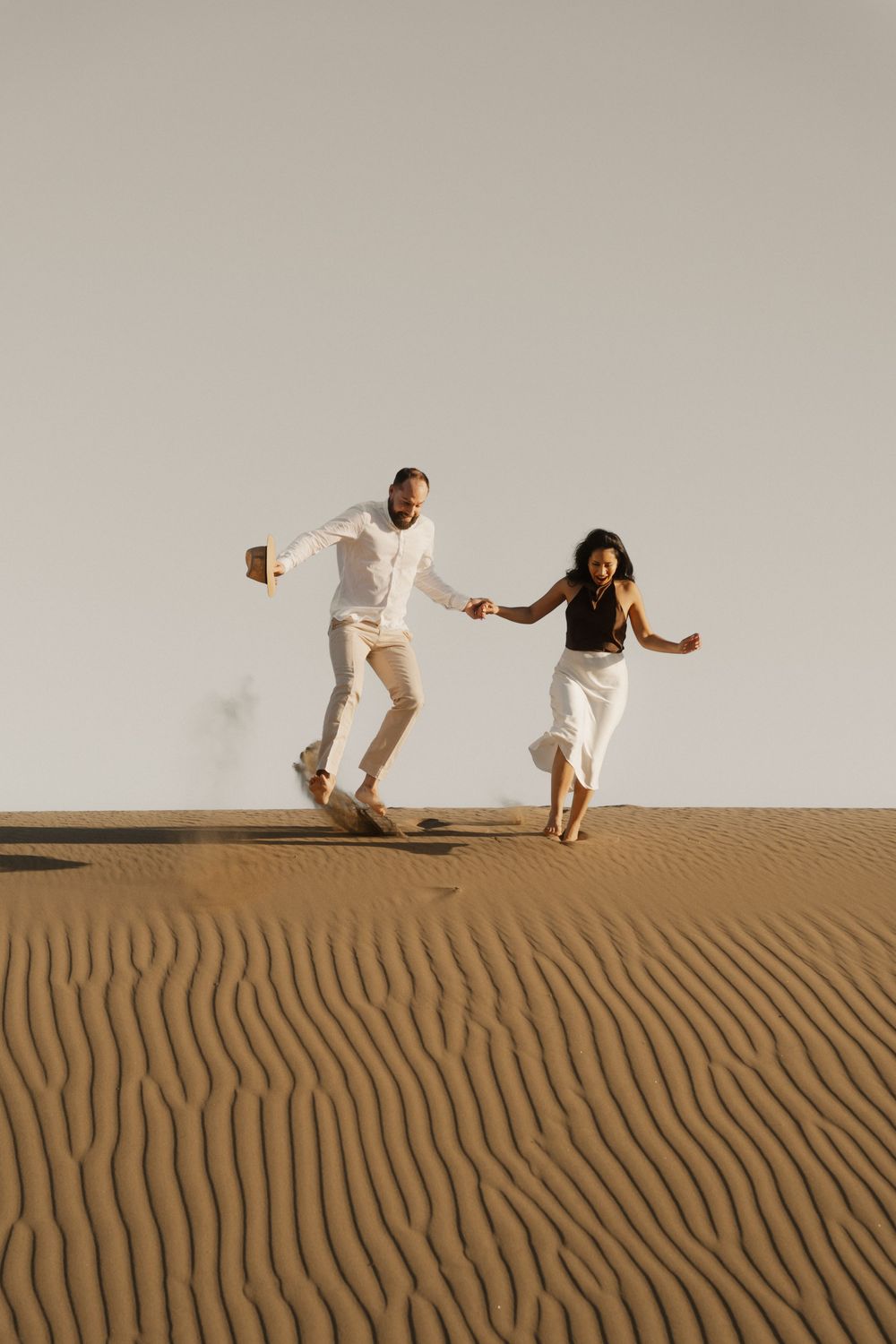 Adventurous couple laughing and walking down a steep sand dune during an editorial photo session.