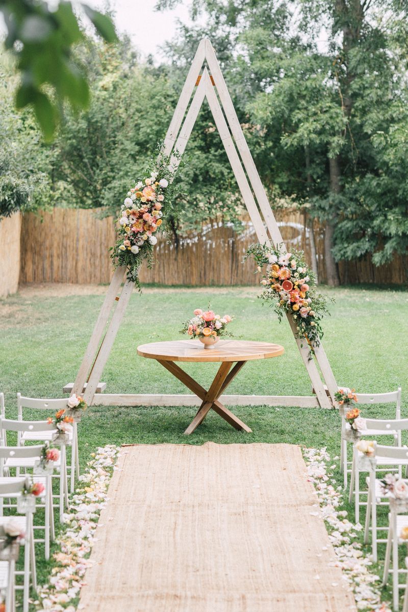 A wooden A-frame wedding arch decorated with coral flowers and white chairs on a grass lawn.