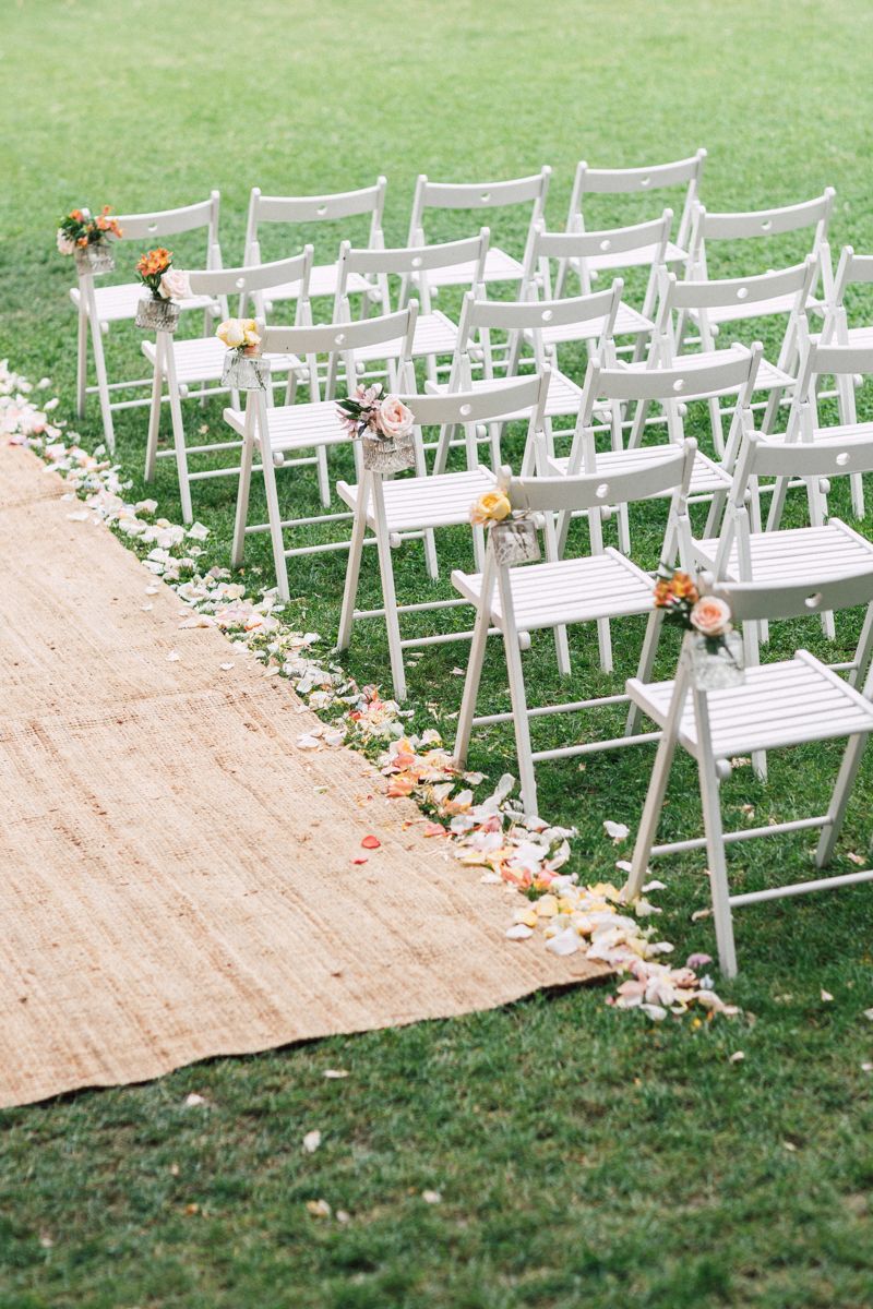 White wooden chairs arranged in rows on grass with a burlap aisle runner decorated with flower petals for an outdoor wedding.