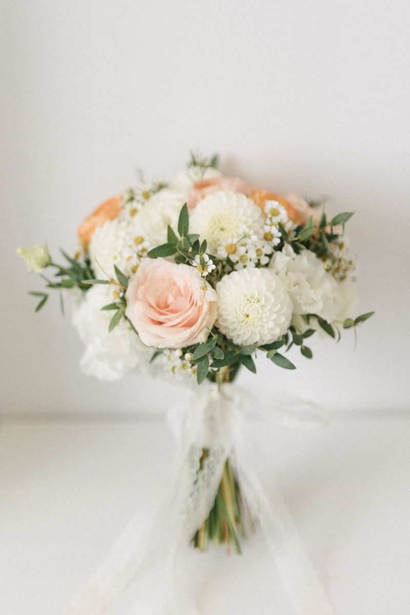 Elegant wedding bouquet with peach roses and white dahlias wrapped in ribbon against white background.
