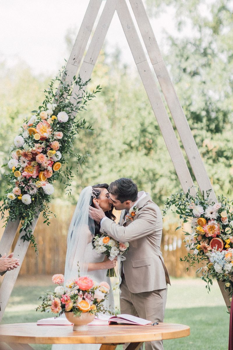 Rustic wooden triangle arch with lush peach and orange floral arrangements at an outdoor wedding.