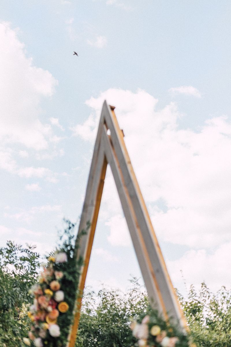 Geometric wooden wedding arch frames against a bright blue sky with wispy clouds.