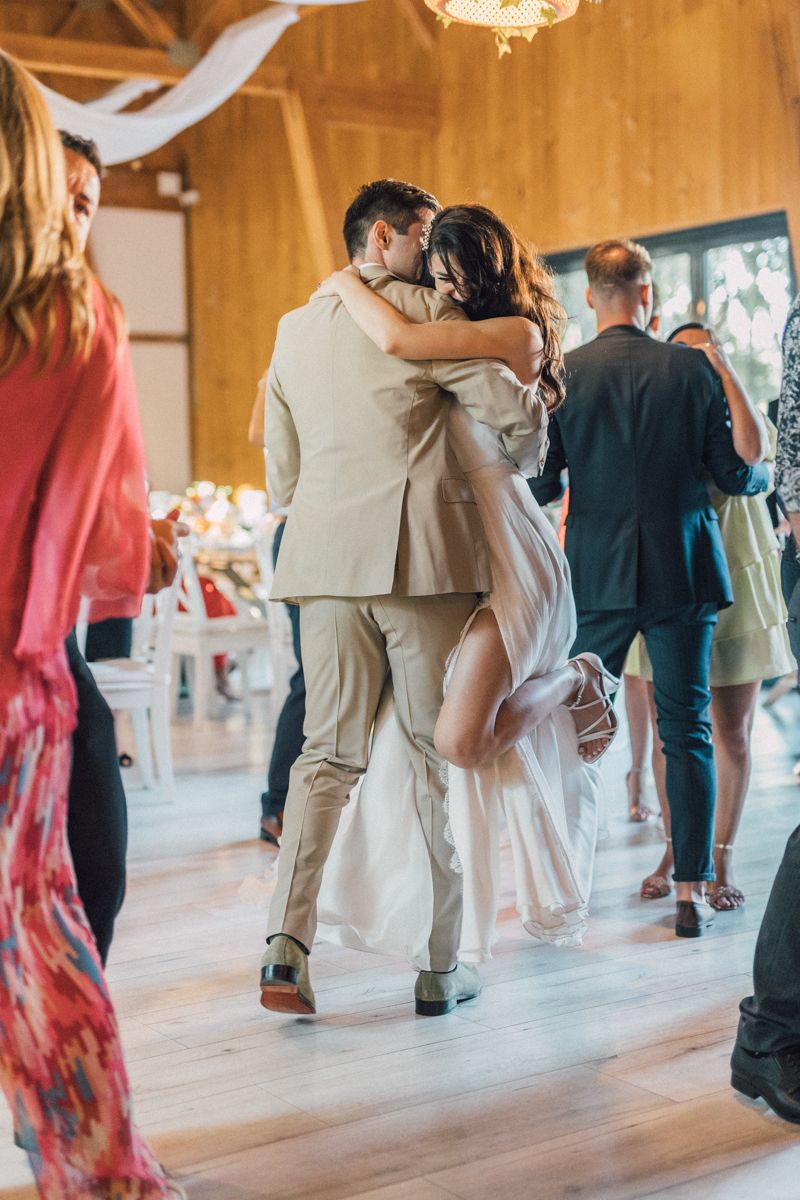 Wedding guests dance in pairs across a sunlit wooden dance floor at a rustic venue.