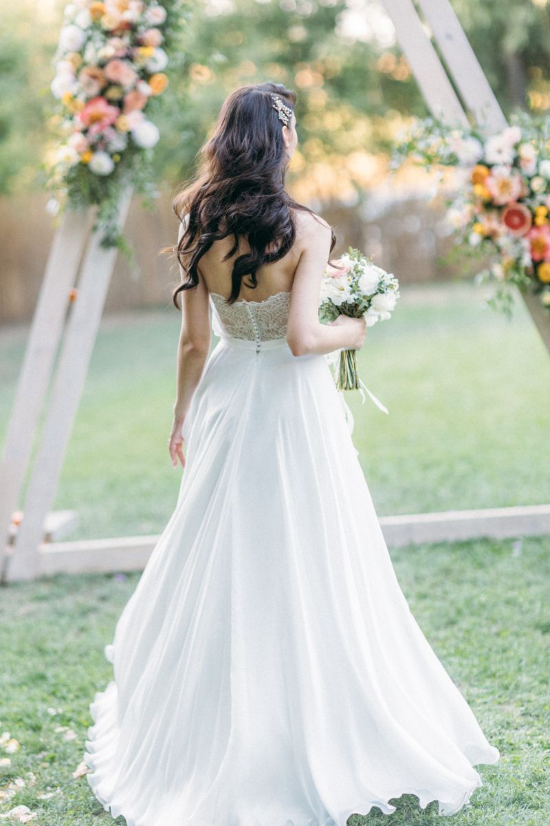 Flowing white wedding dress with floral bouquet against outdoor garden backdrop.