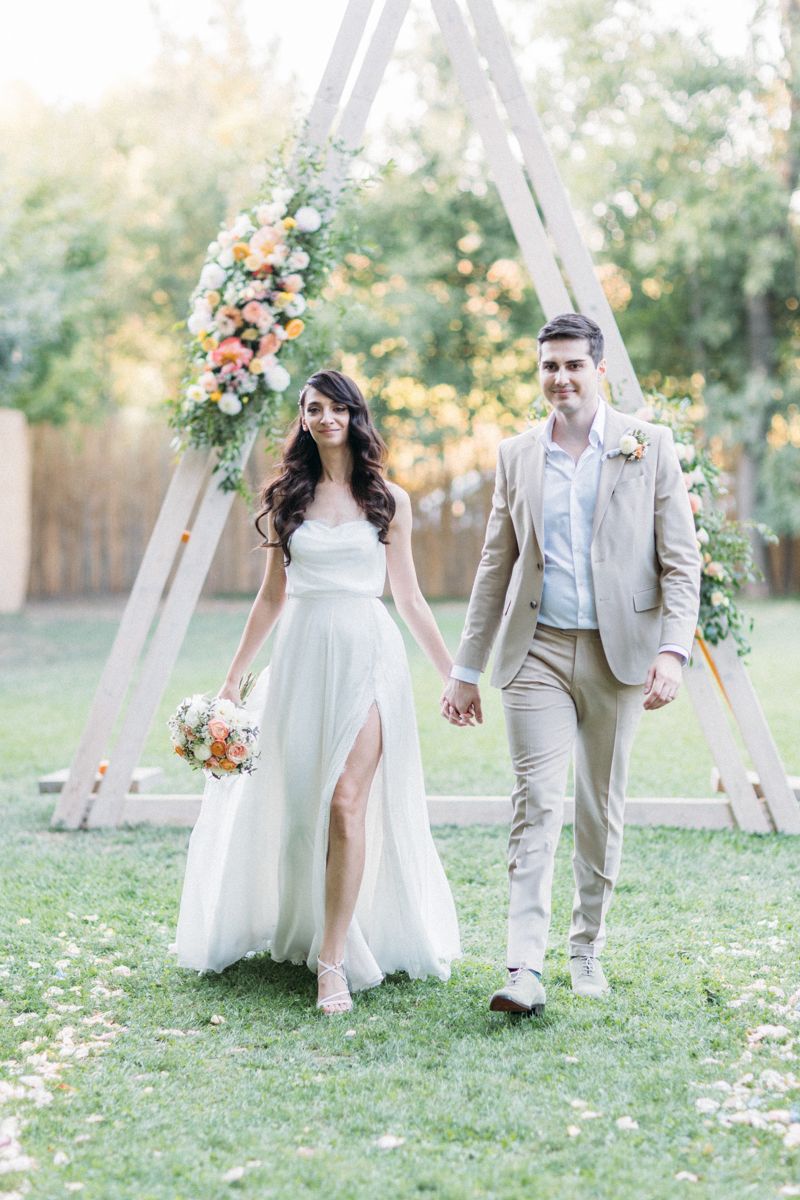 Newlyweds walk hand in hand through garden with white floral arch and scattered petals.