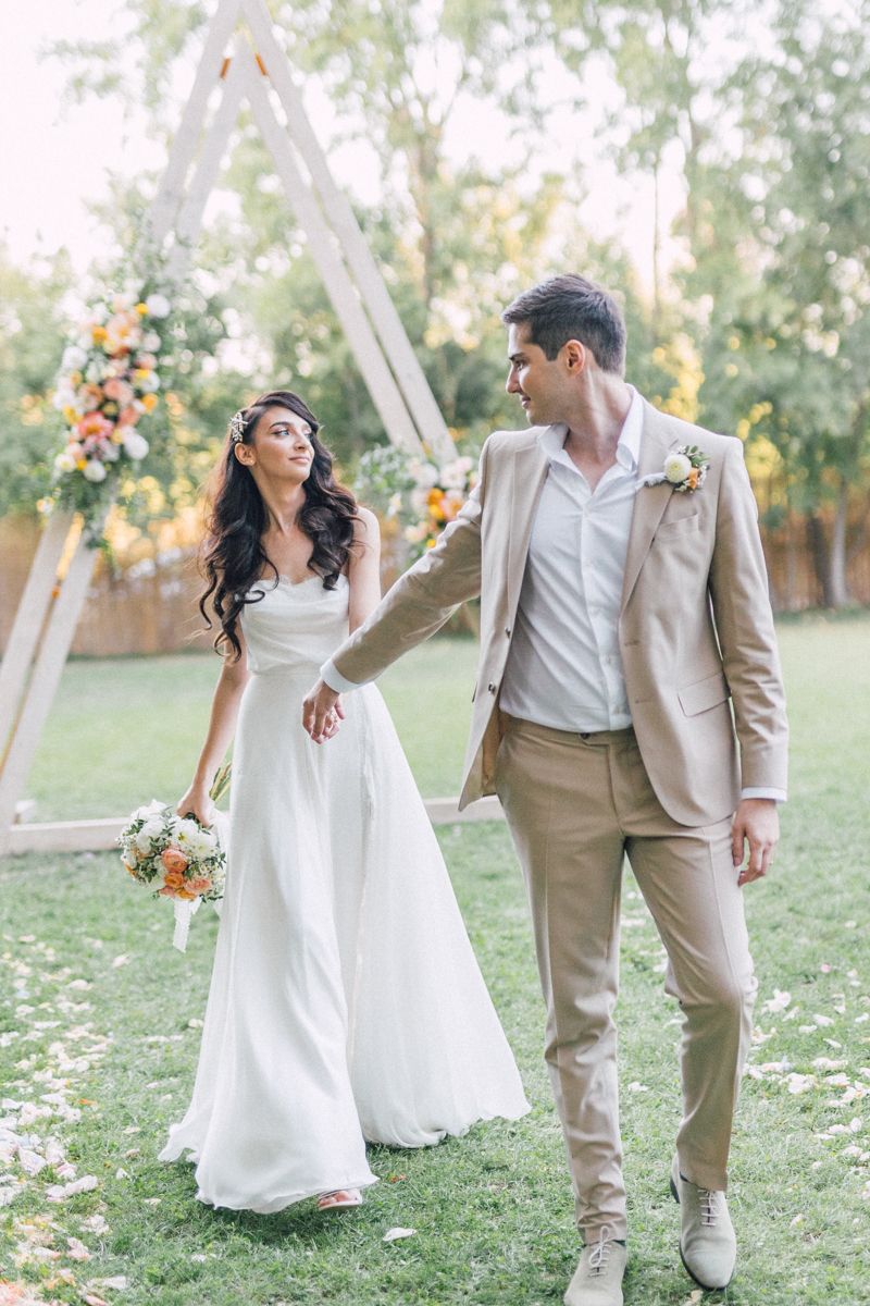 Couple in wedding attire poses playfully on grass with white triangular arch backdrop.