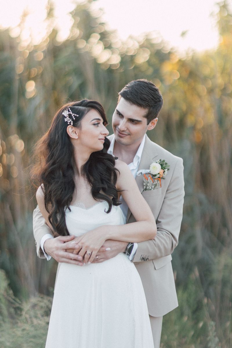 Bride in white dress and groom in beige suit embrace during sunset photo session.