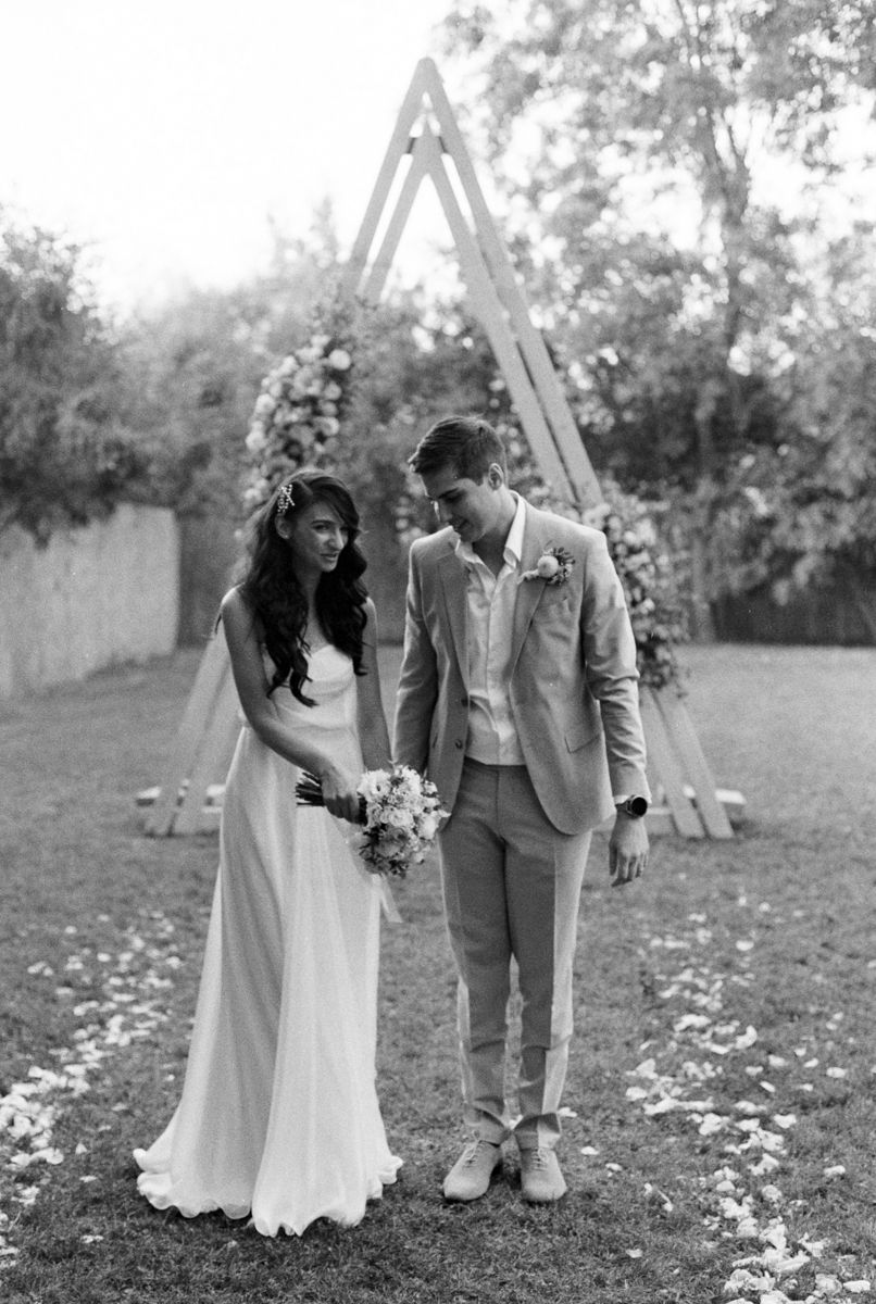 A couple poses together on grass near a wooden ceremony arch with scattered flower petals.