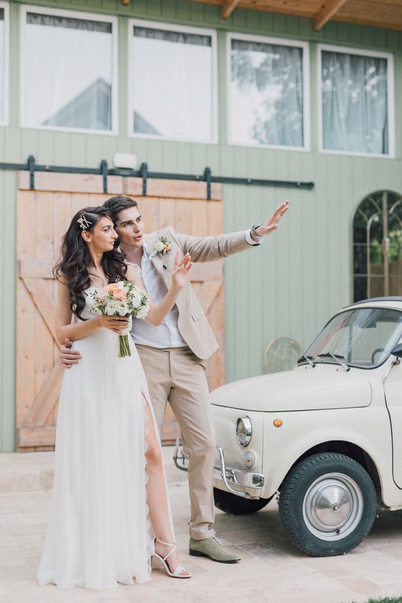 Playful wedding couple posing near vintage white car and rustic green building.