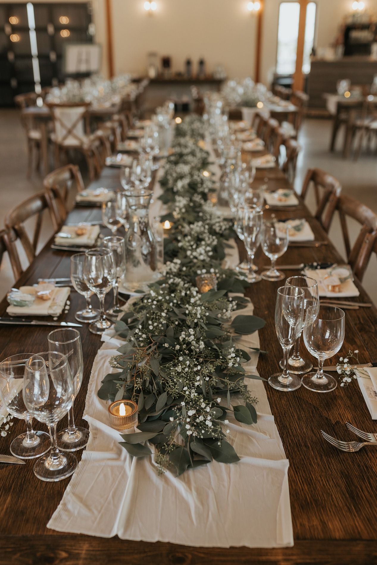 Rustic wedding reception tables set with white linens, greenery centerpieces, wine glasses and wooden cross-back chairs.