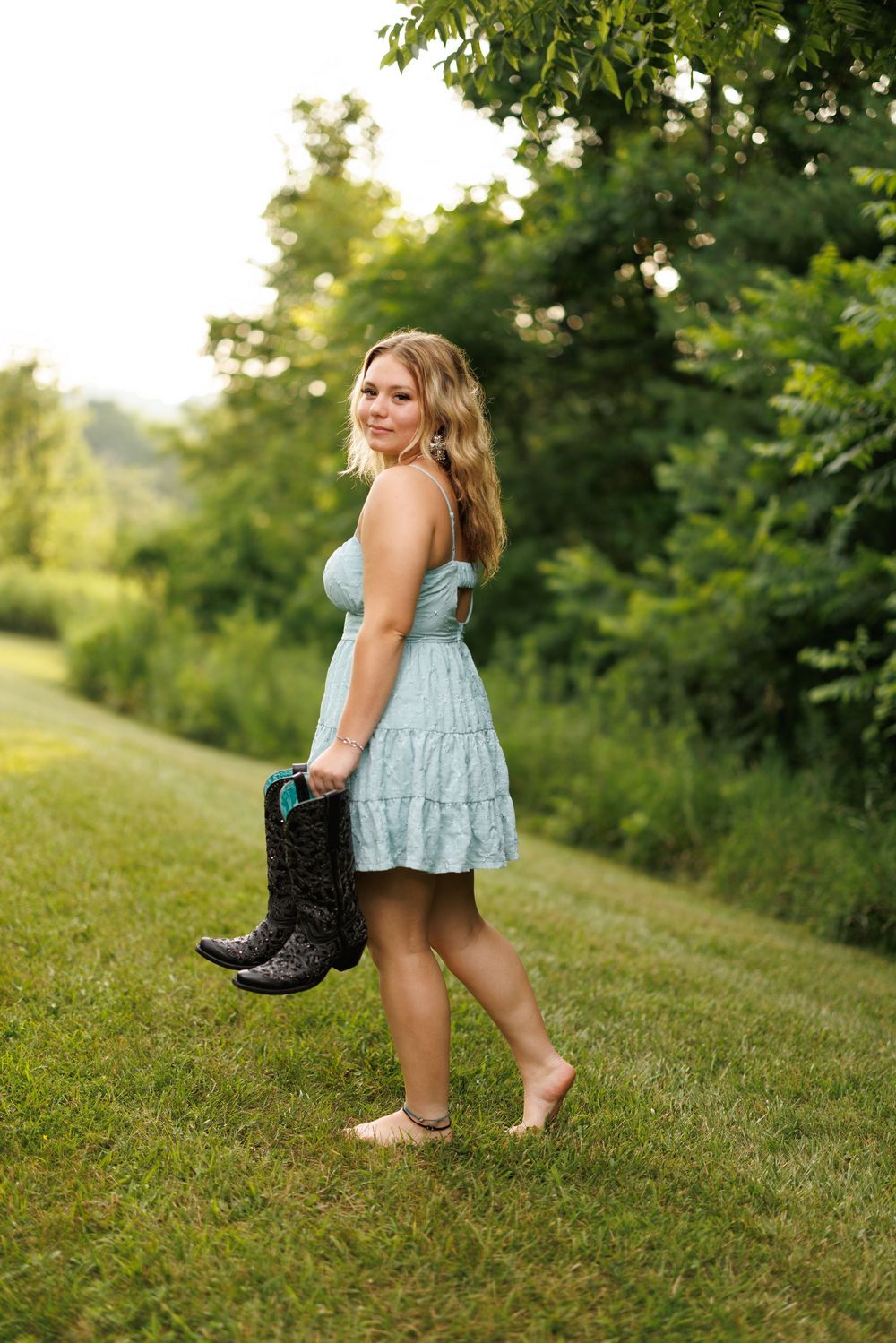 Figure in light blue dress and boots playfully poses in green summer landscape.