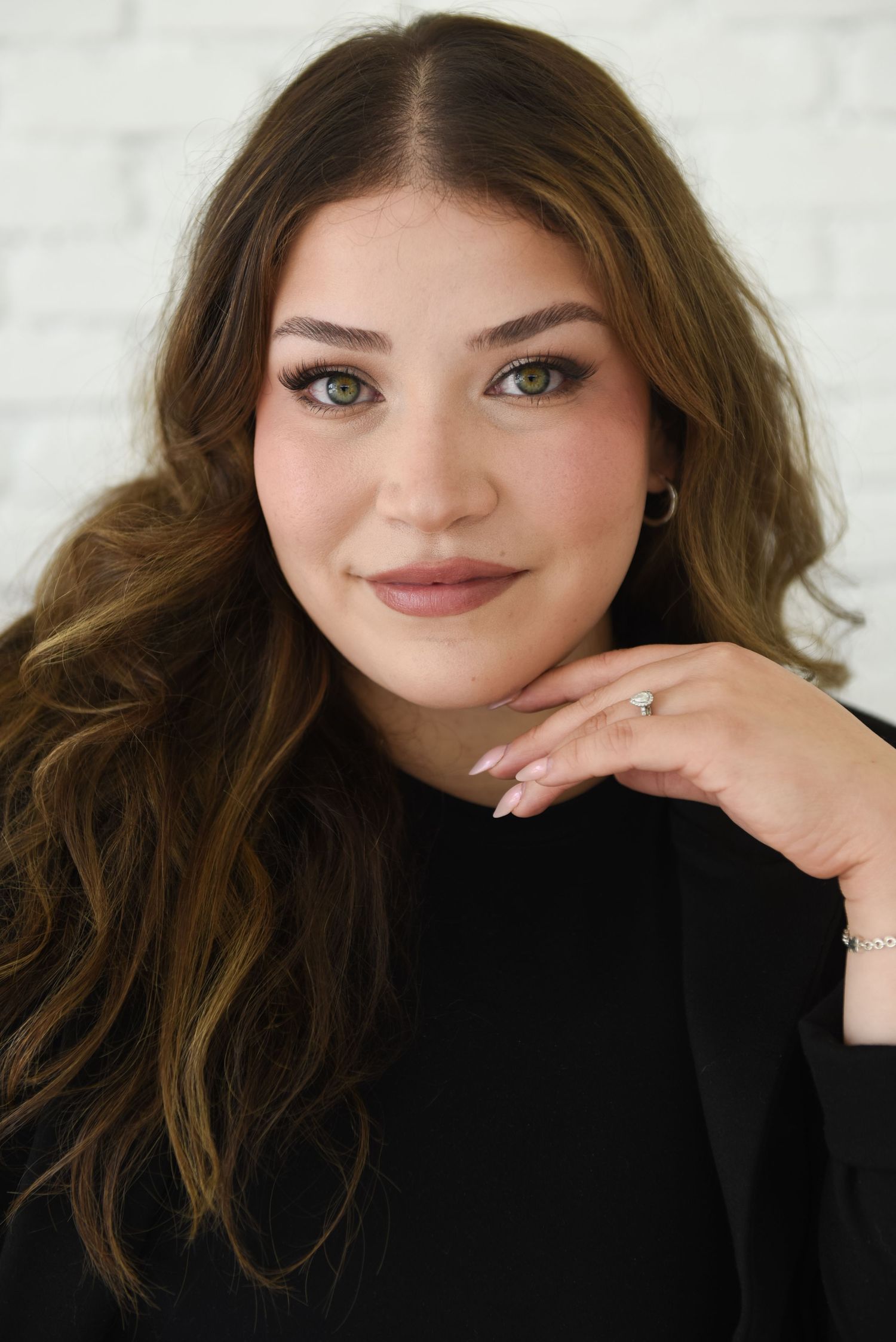 Professional portrait of someone with long wavy brown hair wearing a black top and natural makeup against a light background.