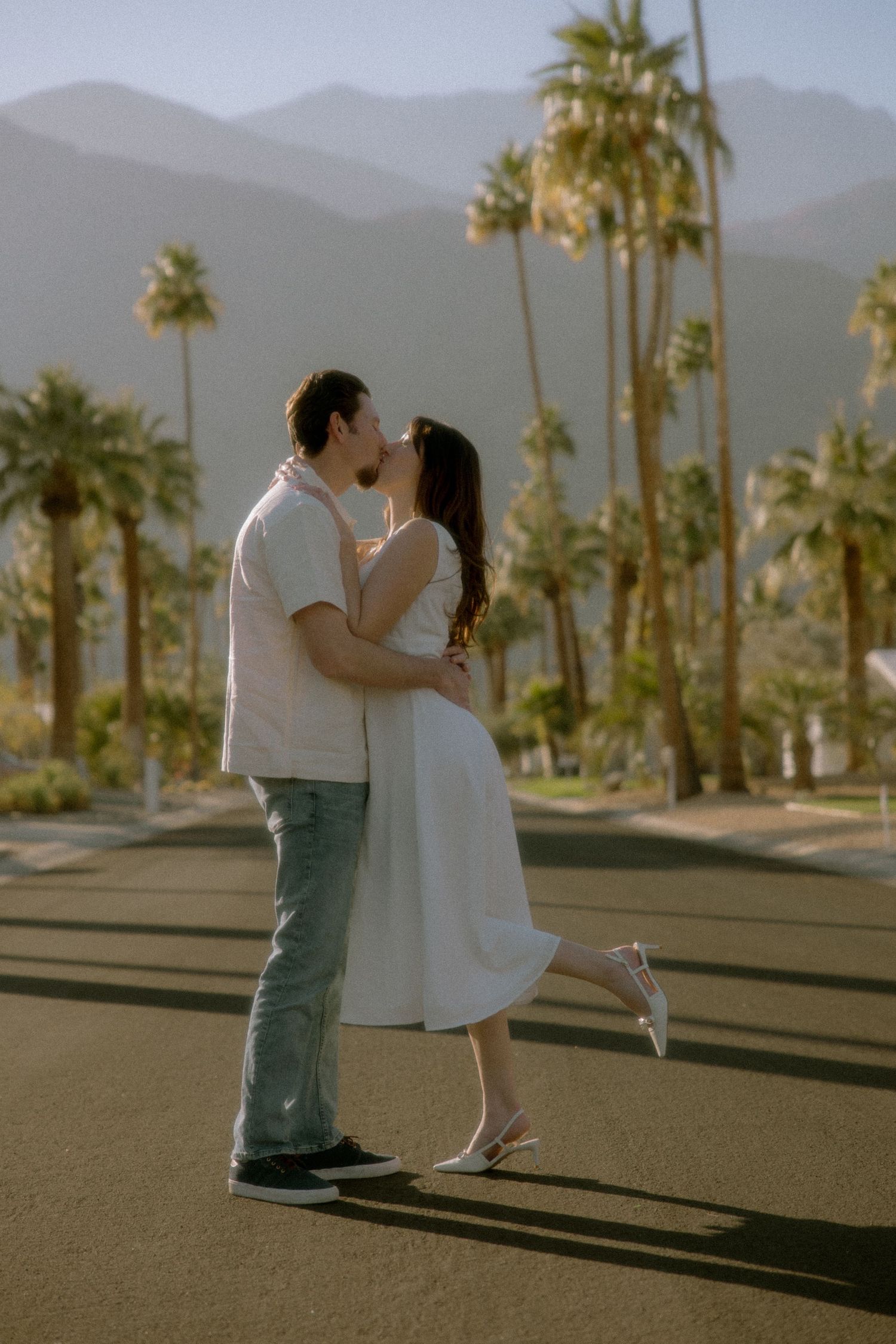 A romantic couple shares a tender kiss against a backdrop of palm trees and mountains in Palm Springs at sunset.