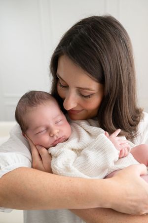 A smiling mother with dark hair gently cradles her sleeping newborn baby wrapped in a white blanket.