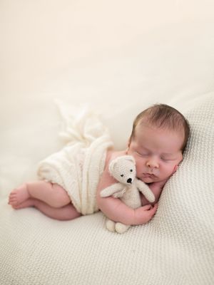 Sleeping newborn baby wrapped in white knit blanket cuddling a small cream teddy bear on a soft white surface.