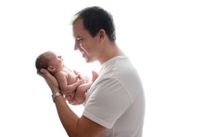 A man in a white t-shirt holds a newborn baby in profile against a light background.