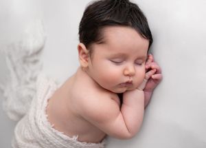 A sleeping newborn baby wearing white lace lies peacefully against a soft white background.