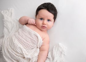A baby wearing a delicate white lace dress lies on soft white fabric in a studio setting.