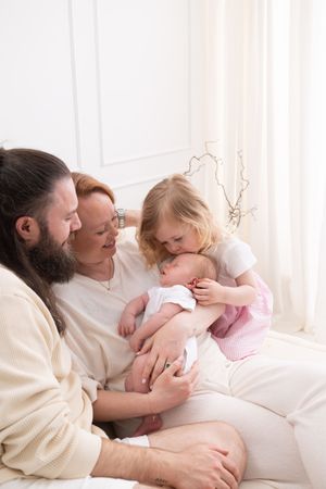 A happy family with a bearded father, redhead mother, toddler girl, and newborn baby sitting together on a white sofa.