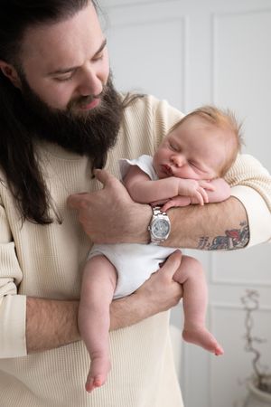 Bearded tattooed father tenderly holds sleeping newborn baby in white onesie against light background.