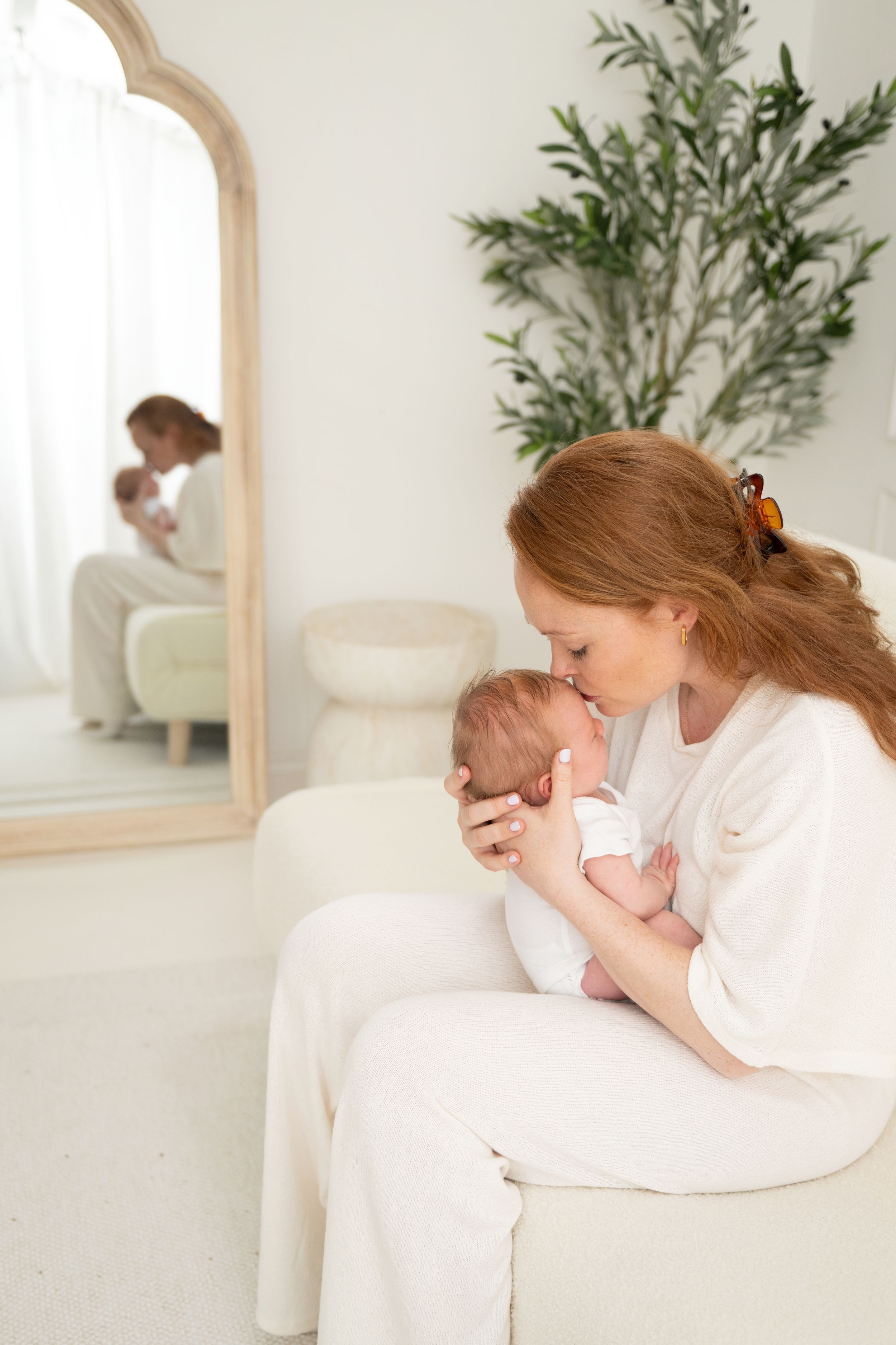 A redheaded mother tenderly kisses her newborn baby while seated on a white chair, with a mirror reflection in the background.