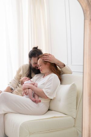 A couple sits on a white chair, tenderly holding their newborn baby in a bright, softly lit room.