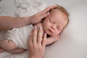 Adult hands gently cradling sleeping newborn baby's face, wearing white knit outfit on soft white backdrop.