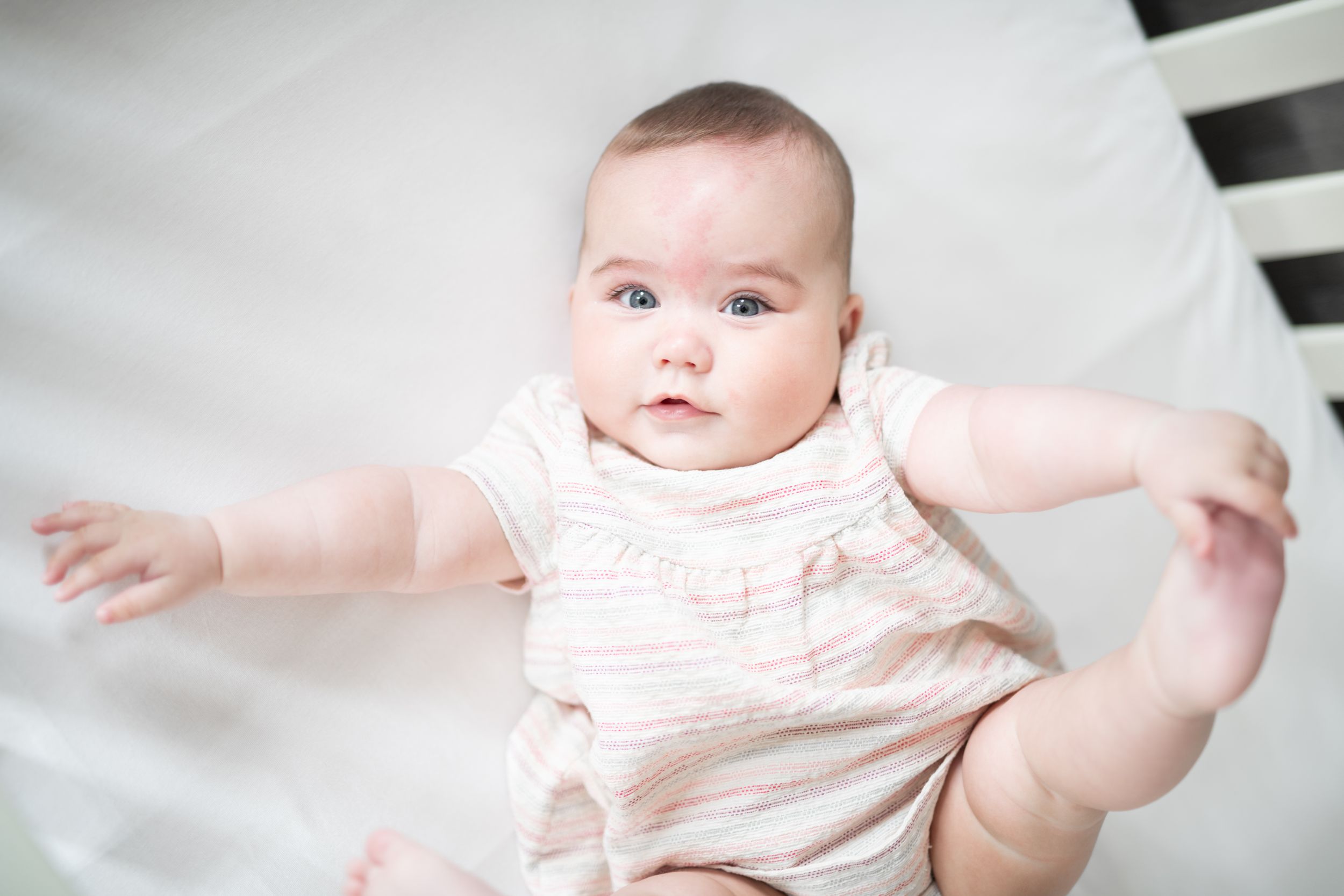 A baby in pink and white stripes smiles and wiggles playfully on a white blanket.