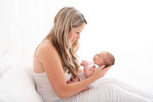 Mother in white dress lying on bed holding and gazing lovingly at newborn baby in natural light setting.