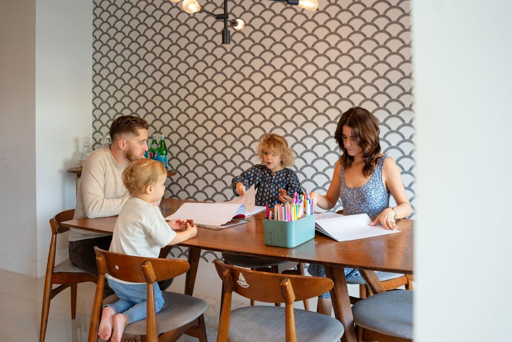 A group sits at a wooden dining table with patterned wallpaper in the background during a casual gathering.