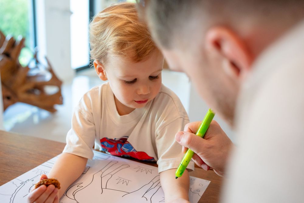 A child practices writing and drawing skills with colorful markers on paper at a wooden table.