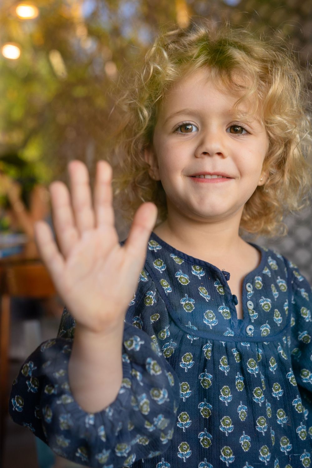 Young child with curly blonde hair wearing a blue patterned shirt waves at the camera with a bright smile.