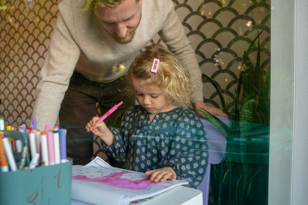 A learning moment captured as someone helps a child with curly hair work on an activity at a desk.