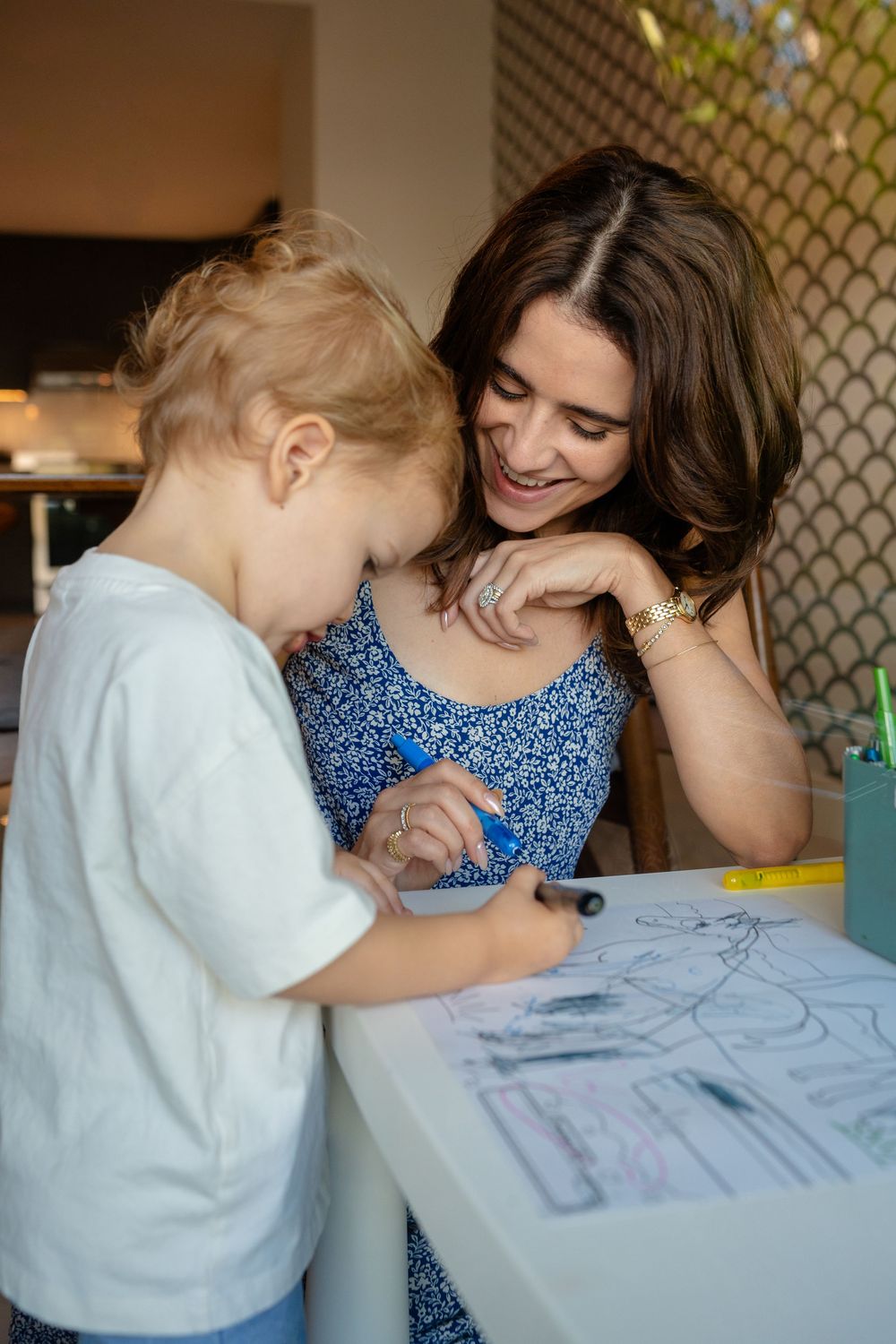 A young child draws on paper at a table while an adult in a blue floral dress watches with interest and encouragement.